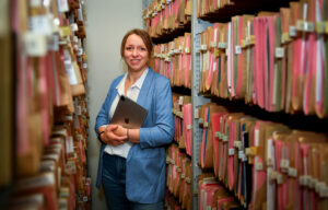 Diana Härter steht mit einem Tablet unter dem Arm zwischen Regalreihen voller Hängeregister.