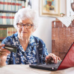 Eine ältere Frau sitzt an einem Laptop und schaut auf eine Kreditkarte.