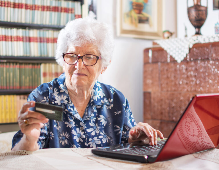 Eine ältere Frau sitzt an einem Laptop und schaut auf eine Kreditkarte.