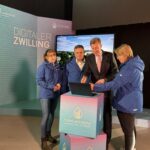 Vier Personen stehen in einem Ausstellungsraum um einen Laptop versammelt und betrachten dessen Bildschirm.