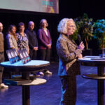 Eine Person spricht auf einer Bühne, im Hintergrund sind die DIGITAL-Awards und mehrere wartende Personen zu sehen.