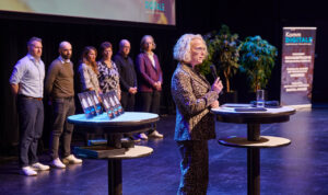 Eine Person spricht auf einer Bühne, im Hintergrund sind die DIGITAL-Awards und mehrere wartende Personen zu sehen.