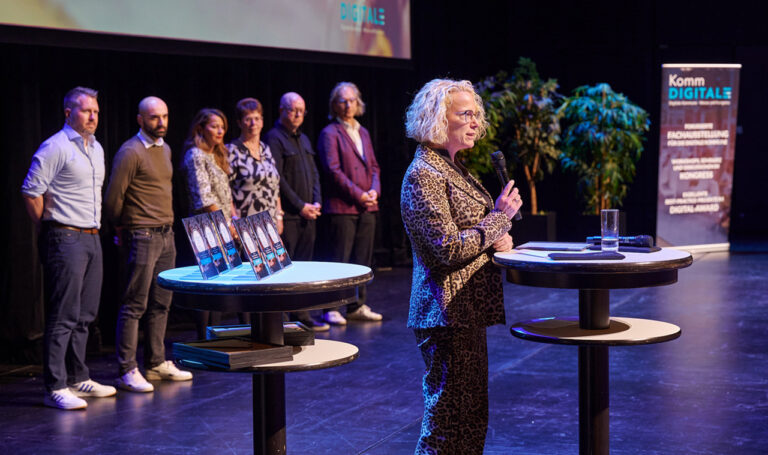 Eine Person spricht auf einer Bühne, im Hintergrund sind die DIGITAL-Awards und mehrere wartende Personen zu sehen.