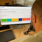 Blick über die Schulter eines Mannes auf einen Monitor, auf dem die farbigen Kacheln des OZG-Dashboard zu sehen sind.