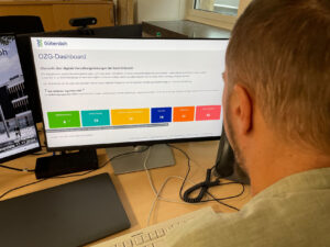 Blick über die Schulter eines Mannes auf einen Monitor, auf dem die farbigen Kacheln des OZG-Dashboard zu sehen sind.