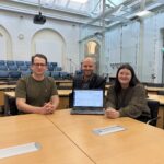 Zu sehen sind 3 Personen die im Großen Sitzungsaal sitzen und einen Laptop präsentieren.