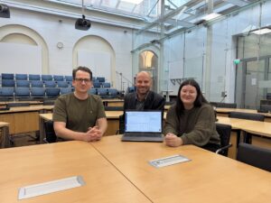 Zu sehen sind 3 Personen die im Großen Sitzungsaal sitzen und einen Laptop präsentieren.