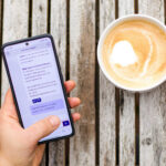 Eine Hand hält ein Smartphone, auf dessen Bildschirm der 115-Chatbot zu sehen ist. Die Hand ruht auf einem Holztisch neben einer Tasse Kaffee.