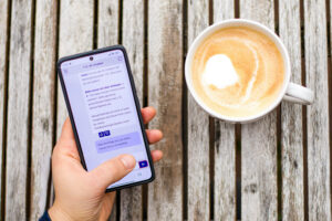 Eine Hand hält ein Smartphone, auf dessen Bildschirm der 115-Chatbot zu sehen ist. Die Hand ruht auf einem Holztisch neben einer Tasse Kaffee.