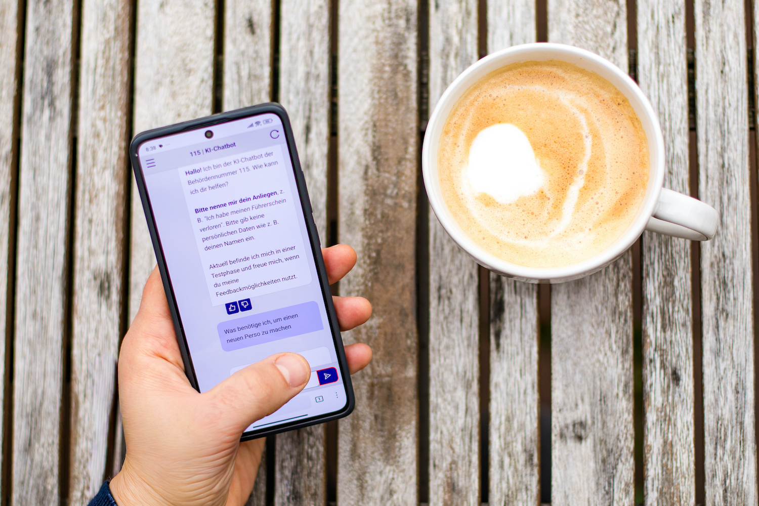 Eine Hand hält ein Smartphone, auf dessen Bildschirm der 115-Chatbot zu sehen ist. Die Hand ruht auf einem Holztisch neben einer Tasse Kaffee.