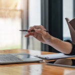 Eine Person sitzt vor einem aufgeklappten Laptop und hat ein Notizbuch in der Hand, dessen Inhalt sie mit den Daten auf dem Laptopbibdschirm vergleicht.