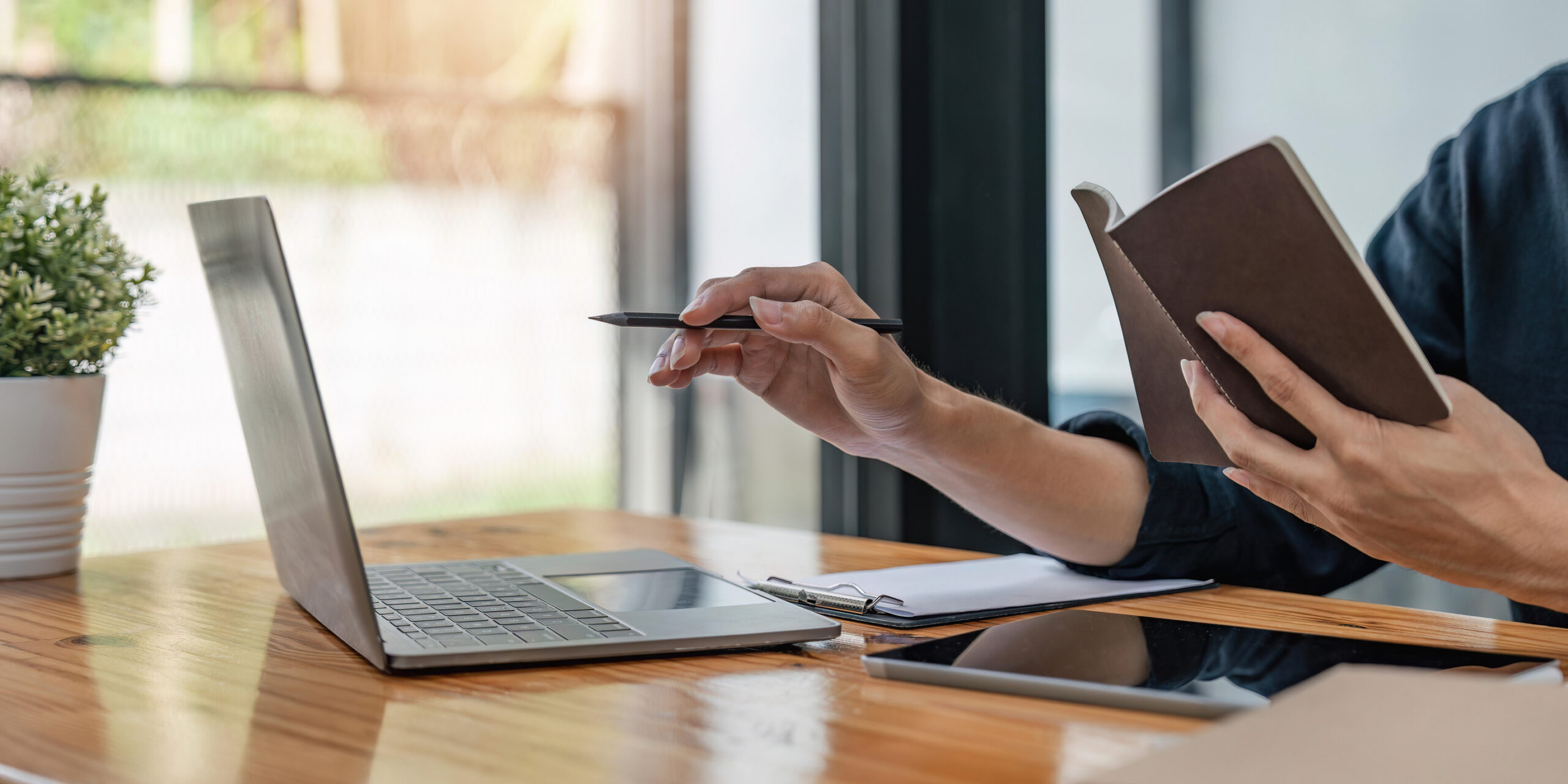 Eine Person sitzt vor einem aufgeklappten Laptop und hat ein Notizbuch in der Hand, dessen Inhalt sie mit den Daten auf dem Laptopbibdschirm vergleicht.