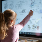 Ein kleines Mädchen zeichnet etwas auf ein Smartboard in einem Klassenzimmer.