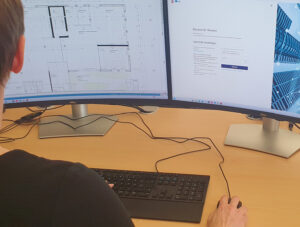 Eine Person sitzt an einem Desktop-Rechner und arbeitet an einem Bauplan.