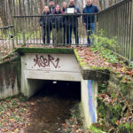 Mehrere Personen stehen auf einer Brücke über einem Bachlauf, eine der Personen hält ein Tablet in der Hand.