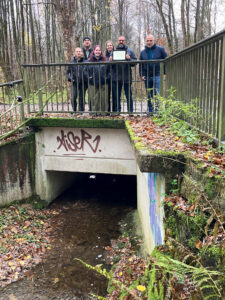 Mehrere Personen stehen auf einer Brücke über einem Bachlauf, eine der Personen hält ein Tablet in der Hand.