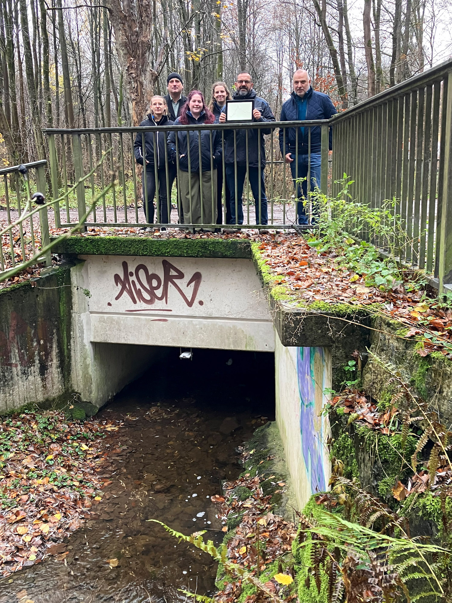 Mehrere Personen stehen auf einer Brücke über einem Bachlauf, eine der Personen hält ein Tablet in der Hand.
