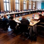 Blick in einen gut besetzten Sitzungssaal mit rechteckiger Tischanordnung.