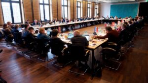 Blick in einen gut besetzten Sitzungssaal mit rechteckiger Tischanordnung.