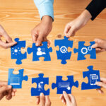 Sieben Hände halten je ein großes Puzzleteil, das mit den anderen Puzzleteilen verbunden werden soll.