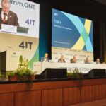 Auf der Bühne einer Verbandsversammlung des Zweckverbands 4IT sitzen mehrere Personen an einem langen Podiumstisch. Links spricht ein Vorsitzender am Rednerpult, hinter ihm ist sein Bild auf einer großen Leinwand zu sehen. Rechts zeigt die Präsentation den Tagesordnungspunkt „TOP 6 – Statusbericht Expertenkreise“. Vor der Bühne stehen Blumengestecke und Pflanzen.