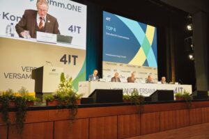 Auf der Bühne einer Verbandsversammlung des Zweckverbands 4IT sitzen mehrere Personen an einem langen Podiumstisch. Links spricht ein Vorsitzender am Rednerpult, hinter ihm ist sein Bild auf einer großen Leinwand zu sehen. Rechts zeigt die Präsentation den Tagesordnungspunkt „TOP 6 – Statusbericht Expertenkreise“. Vor der Bühne stehen Blumengestecke und Pflanzen.