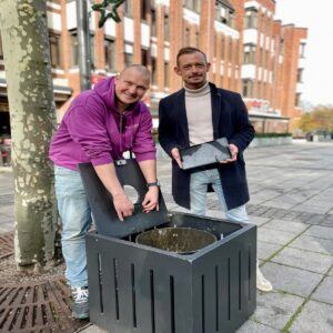 Dr. Daniel Trauth und Christian Henicke präsentieren Füllstandsensor und Tablet neben einem öffentlichen Abfallbehältnis in Grevenbroich.