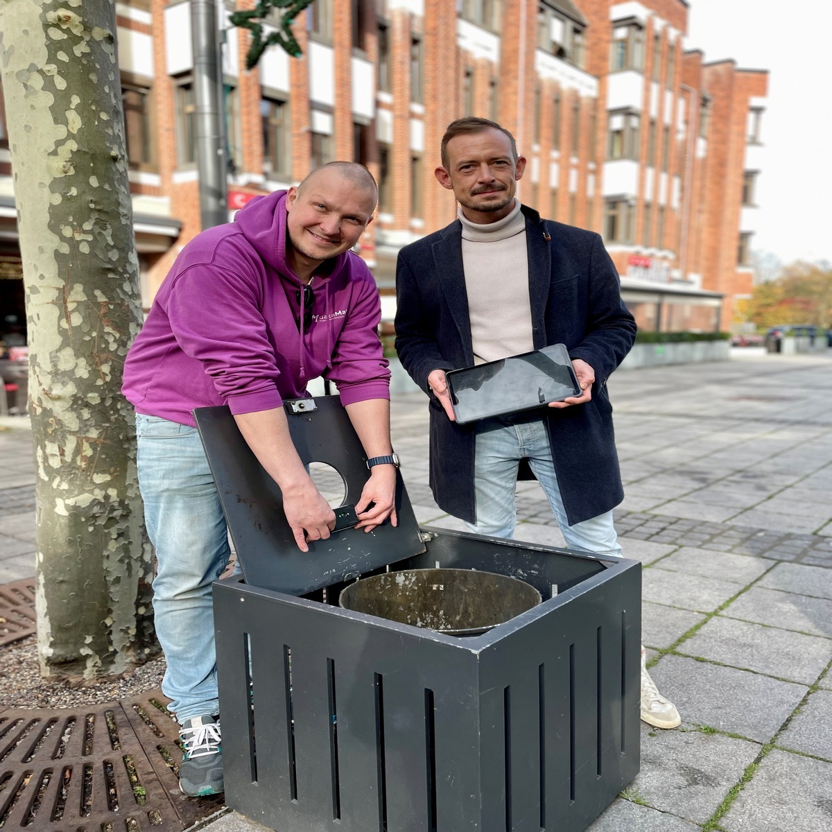 Dr. Daniel Trauth und Christian Henicke präsentieren Füllstandsensor und Tablet neben einem öffentlichen Abfallbehältnis in Grevenbroich.