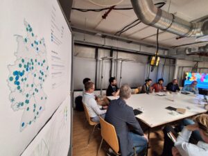 Mehrere Personen sitzen an einem Projekttisch zusammen, hinter ihnen ist eine Deutschlandkarte mit Markierungen zu sehen.