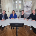 Zwei Männer und zwei Frauen sitzen nebeneinander an einem rundlichen Tisch und unterzeichnen Unterlagen.