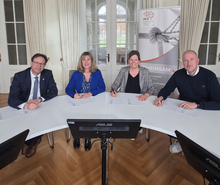 Zwei Männer und zwei Frauen sitzen nebeneinander an einem rundlichen Tisch und unterzeichnen Unterlagen.