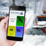 Eine Person hält ein Smartphone in der Hand auf dem eine AIDA-ORGA-App zu sehen ist. Im Hintergrund ist ein Schneepflug zu sehen, der in einer verschneiten Kommune unterwegs ist.