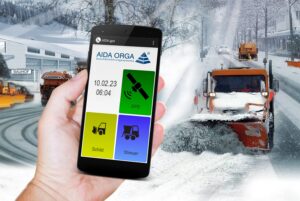 Eine Person hält ein Smartphone in der Hand auf dem eine AIDA-ORGA-App zu sehen ist. Im Hintergrund ist ein Schneepflug zu sehen, der in einer verschneiten Kommune unterwegs ist.