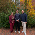 Drei Personen stehen nebeneinander im Freien neben einem Gebäude, im Hintergrund sind vom Herbst gezeichnete Sträucher zu sehen.