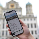 Ein Smartphone mit einer Stadtansicht von Augsburg im Hintergrund, auf dem Bildschirm des Smartphones ist die Stadt Augsburg-App zu sehen.