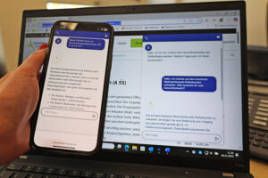 Eine Person hält ein Smartphone in der Hand, auf dem der Chatbot des Gesundheitsamts der StädteRegion Aachen zu sehen ist. Im Hintergrund steht ein Laptop auf dem ebenfalls der Chatbot angezeigt wird.