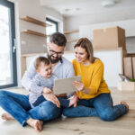 Junge Familie mit Kleinkind und Tablet vor Umzugskartons im neuen Zuhause