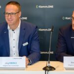 Zwei Vertreter von Komm.ONE sitzen nebeneinander bei einer Pressekonferenz. Links spricht Jörg Eberle lächelnd ins Mikrofon, rechts hört Bernd Krakau aufmerksam zu. Beide tragen dunkle Anzüge und sitzen vor einer Komm.ONE-Hinterwand.