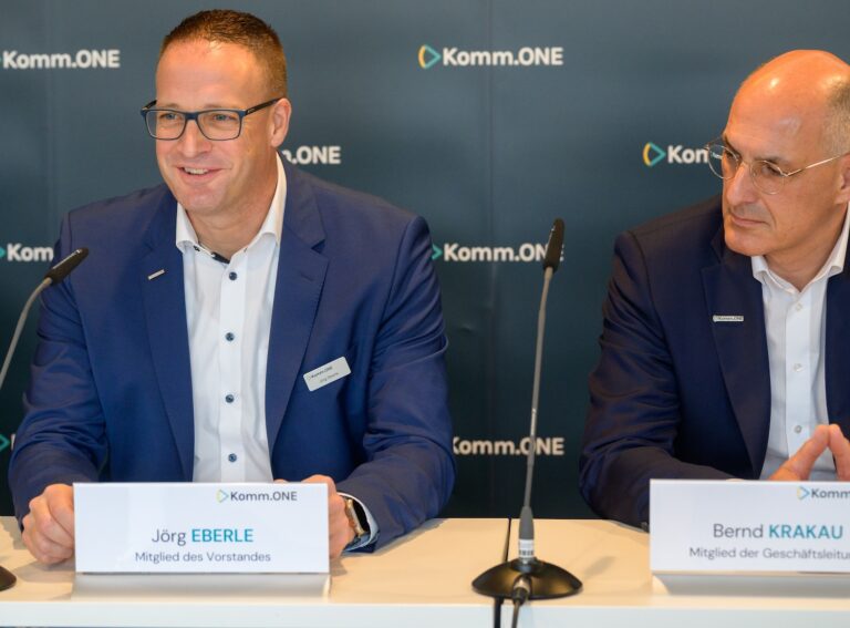 Zwei Vertreter von Komm.ONE sitzen nebeneinander bei einer Pressekonferenz. Links spricht Jörg Eberle lächelnd ins Mikrofon, rechts hört Bernd Krakau aufmerksam zu. Beide tragen dunkle Anzüge und sitzen vor einer Komm.ONE-Hinterwand.