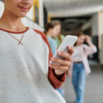 Eine Schülerin hält ein Smartphone in der Hand, im Hintergrund ist verschwommen ein Schulflur mit weiteren Schülerinnen und Schülern zu sehen.