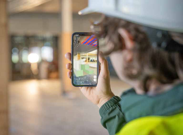Eine Person mit Bauhelm hält ein Smartphone in der Hand auf dem die 3D_Darstellung eines Innenraums zu sehen ist.