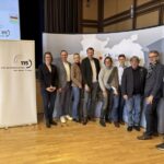 Mehrere Personen stehen nebeneinander in einem Raum, im Hintergrund ist ein Aussteller zu sehen, der die 115 abbildet.