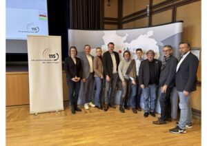 Mehrere Personen stehen nebeneinander in einem Raum, im Hintergrund ist ein Aussteller zu sehen, der die 115 abbildet.