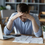 Junger Mann sitzt mit Laptop, Taschenrechner und einigen Papieren in sorgenvoller Haltung am Schreibtisch.