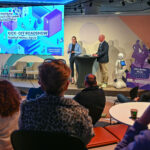 Abgedunkelter Saal mit Publikum, im Bildhintergrund eine helle Bühne mit Projektion "Kick-off Roadshow Hanau", zwei Personen auf der Bühne