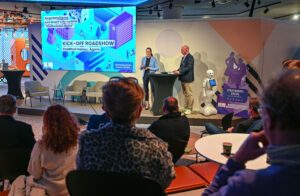 Abgedunkelter Saal mit Publikum, im Bildhintergrund eine helle Bühne mit Projektion "Kick-off Roadshow Hanau", zwei Personen auf der Bühne