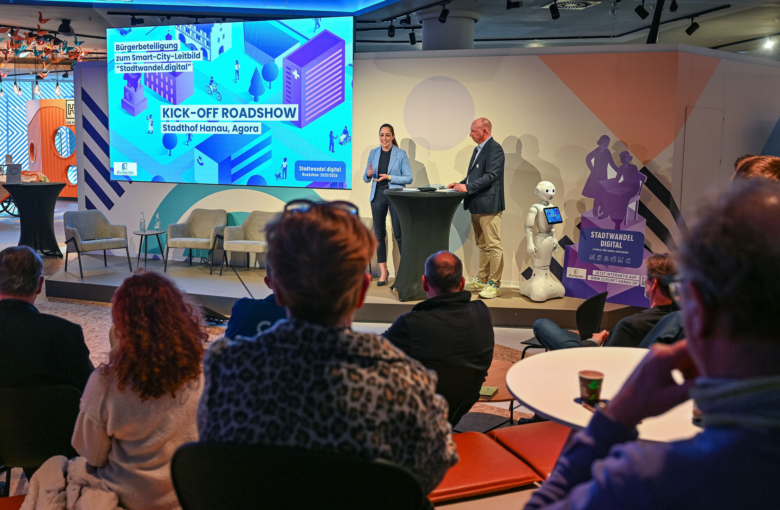 Abgedunkelter Saal mit Publikum, im Bildhintergrund eine helle Bühne mit Projektion "Kick-off Roadshow Hanau", zwei Personen auf der Bühne