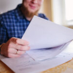 Mann am Schreibtisch mit Laptop, hantiert mit Papieren.