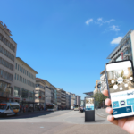 Die Pforzheimer Innenstadt. Im Vordergrund hebt eine Hand ein Handy, im Hintergrund ist eine Informationsstele zu sehen