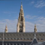 Blick vom Dach auf das Wiener Rathaus: neogotischer Bau mit zentralem Turm, Uhr und filigranen Türmchen vor blauem Himmel.